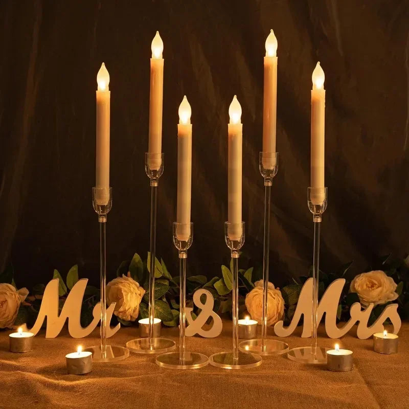 Tall lit candles in candletown.net acrylic holders are on a table with scattered white roses and green leaves. Wooden Mr & Mrs letters and small tea lights create a warm, romantic ambiance against a dark background, blending traditional charm with a modern aesthetic.