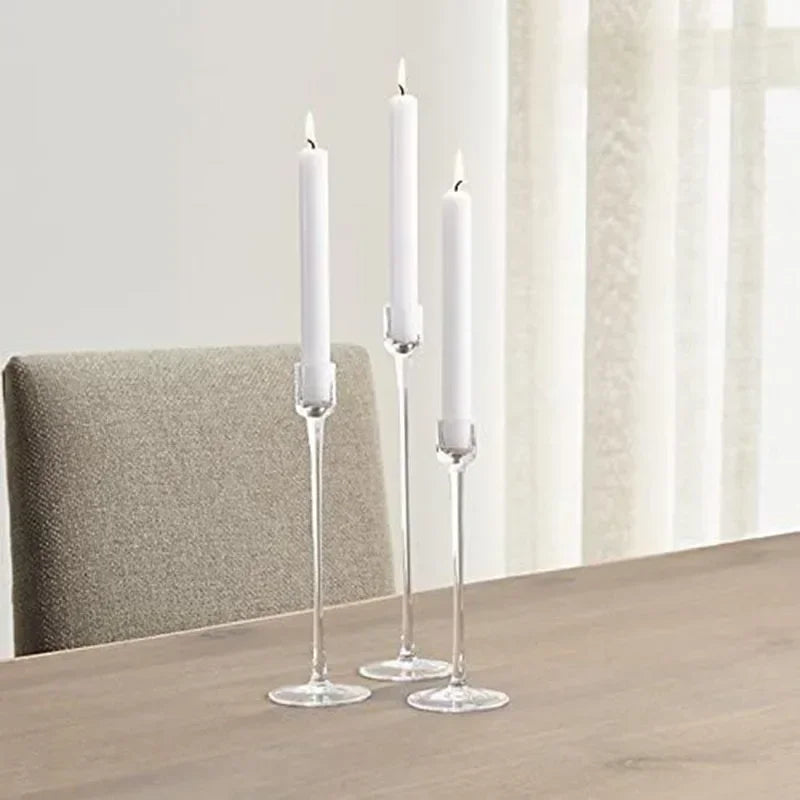 Three tall white candles in clear acrylic holders from candletown.net sit staggered on a wooden table, enhancing the modern look against a light curtain and beige upholstered chair. Ideal for elevating your decor.