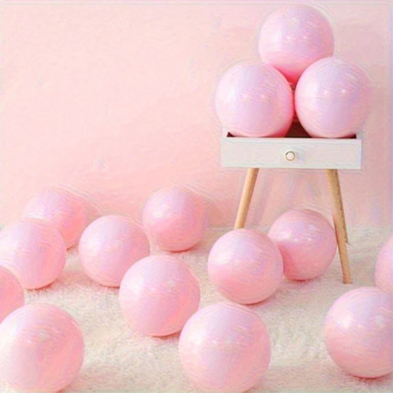 The image features an assortment of pink latex balloons from candletown.net, dispersed over a fluffy white carpet. Several balloons are placed on a small three-legged wooden stool, set against a soft pink background, creating an inviting party decoration scene.